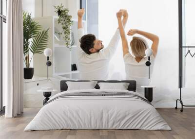 Back view of caucasian couple in casual attire stretching their limbs after night sleep while in bed of bright bedroom. Relaxed family man and woman starting new sunny day in modern flat. Wall mural