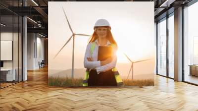 Beautiful caucasian woman in white helmet working with digital tablet at renewable energy farm. Female inspector controlling functioning of wind turbines outdoors. Wall mural