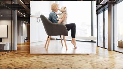 Beautiful slim woman looking at cute dog nose to nose while staying in armchair with feet standing on toes. Playful short-haired female keeper laughing together with canine buddy on weekend at home. Wall mural