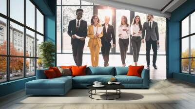 Business People Walking Toward Camera. Full length of group of happy young business people in formal wear having work meeting in modern office with large window Wall mural