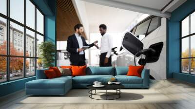 Car salesman discusses electric SUV with businessman. Charging station and modern vehicle highlight eco-friendly options. Professional interaction emphasizes business deal in automotive industry. Wall mural