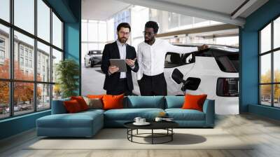 Car salesman using tablet to explain features of electric SUV to potential buyer in modern dealership. Business meeting focuses on environmentally friendly transportation and automotive technology Wall mural