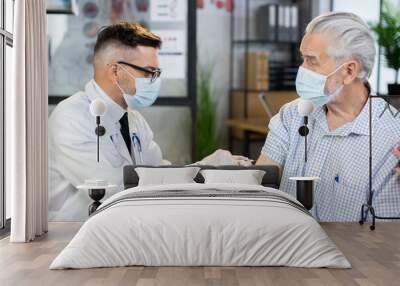 Caucasian male doctor in face mask and gloves doing injection for aged patient at modern clinic. Senior man getting vaccinated for protection from virus. Health care insurance. Wall mural