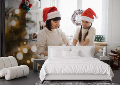 Caucasian mother and daughter wearing Santa hats knead dough in cozy kitchen. Scene filled with Christmas decorations and holiday cheer, capturing family bonding and joyful preparation. Wall mural