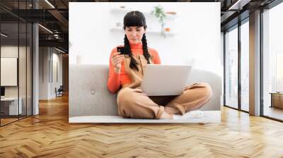 Caucasian woman in casual attire sits on sofa using laptop for online shopping. She holds credit card, making digital transaction. Bright modern living room setting conveys convenience and technology. Wall mural