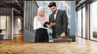 Charming woman in hijab with digital tablet and muslim man with smartphone standing together near office building. Two colleagues using modern gadgets for work outdoors. Wall mural