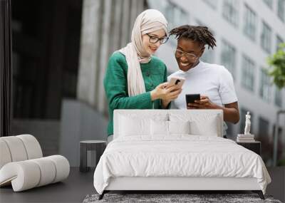 Charming woman in hijab withsmartphone and african female with phone standing together near office building. Two women colleagues using modern gadgets for work outdoors. Wall mural