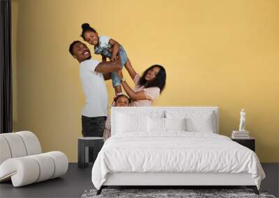 Cheerful african man holding excited daughter on hands. Woman and another daughter looking posing at camera, standing isolated over yellow studio background, panorama, banner, free copy space Wall mural