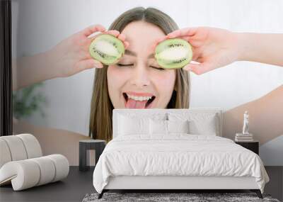 Cheerful beautiful young woman holding two cut kiwis on her eyes for fun joke, smiling and showing tip of her tongue, indoors studio shoot. Beauty and skin care concept Wall mural