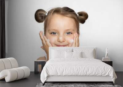 Child face skin care and protection, cold cream. Portrait of cute little caucasian 3 years old girl, applying winter cream, posing on white studio background and smiling Wall mural