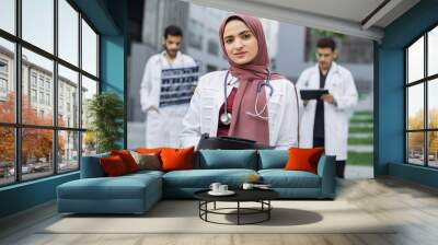 Close up of female Muslim pleasant doctor in hijab, standing with clipboard outdoors in front of hospital building, while two handsome Arabian men doctors working with x-ray and tablet pc behind Wall mural