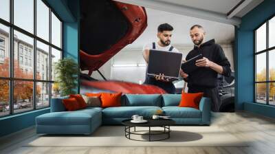 Close up of two young bearded engineer mechanic under car hood checking engine with laptop and tablet using electronic diagnostic equipment to tune sport red vehicle. Wall mural