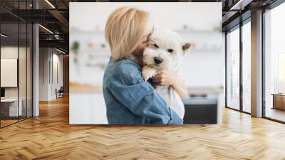 Close up view of tender young woman cuddling little white dog while holding clever pet in arms indoors. Caring short-haired lady in jeans outfit getting great bonding opportunity via canine hugs. Wall mural