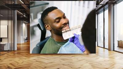 Closeup of a young african man with beautiful smile at the dentist. Dental clinic. Dentist makes comparison of the patient teeth with the dental whitening chart. Wall mural