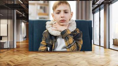 Cold and flu season. Sick teenage boy with high fever and sore throat and feeling bad, sitting at home on the sofa. Little child touches his throat with his hand feeling pain. Wall mural