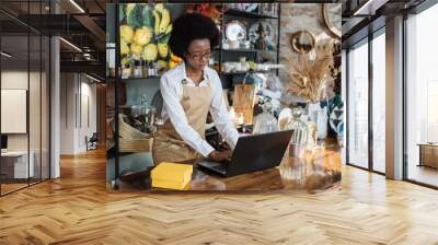 Competent saleswoman in beige apron using wireless laptop while working at shop with various decor. African woman sitting at counter and using modern gadget. Wall mural