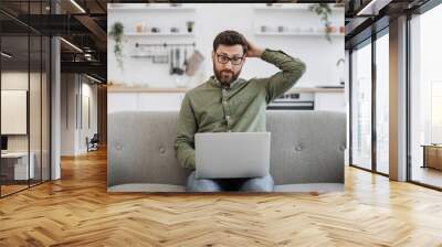 Confused bearded male wearing eyeglasses and casual attire shrugging shoulders while sitting on couch with modern laptop and looking at camera. Concept of doubt, people and technology. Wall mural