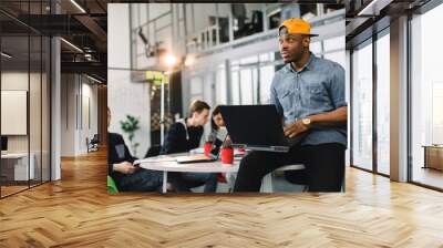 Dark skinned African american coworker in jeans shirt and yellow cap and using laptop in modern office. Multiracial young creative people are working together with laptop, tablet, smart phone. Wall mural