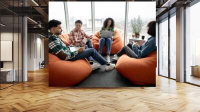 Diverse team of managers in casual wear having conversation while resting on cozy poufs in creative office. Confident collaborators improving corporate strategy using modern technologies in workplace. Wall mural