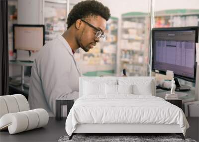 Experienced African American male pharmacist working on pc in modern pharmacy and writing working notes Wall mural