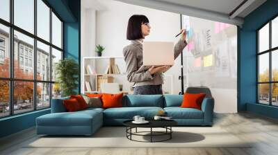 Female administrative manager preparing for presentation at work. Caucasian woman in glasses holding laptop pc and looking on financial graphic on glass wall before office meeting. Wall mural