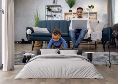 Focused male freelancer sitting on sofa and using modern laptop for remote work. Pretty boy playing with various toys while father working. Wall mural