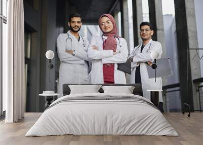 Front view of high-skilled team of three young confident Saudi Arab doctors, standing outside the hospital and looking at camera. Pretty Muslim lady in hijab stands in front of her two male colleagues Wall mural