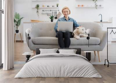 Full length view of barefoot woman looking at computer screen while touching white terrier on top of head in kitchen. Joyful female adult making internet researches while utilizing laptop at home. Wall mural