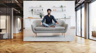 Full length view of mature person doing yoga exercise while sitting with legs crossed on couch in dining room. Barefoot indian man in spectacles relaxing with closed eyes after working day at home. Wall mural