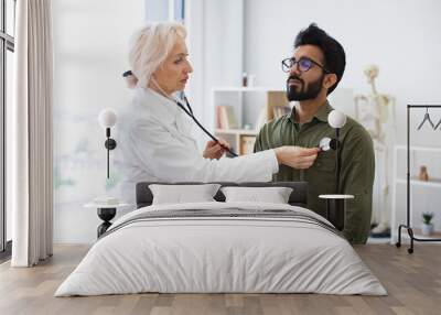 General practitioner examining heartbeat while providing regular checkup in clinic. Side view of focused elderly lady in white coat using stethoscope while young man sitting on exam couch. Wall mural