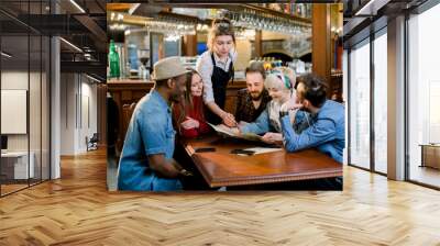 Group of diverse young friends sitting together at a table in a trendy fast food restaurant ordering food from a waitress Wall mural