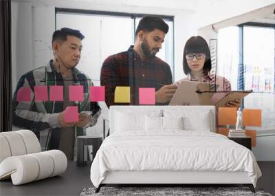 Group of young professionals collaborating on project in modern office. Team gathered in front of glass wall with colorful sticky notes while using digital devices for brainstorming and planning. Wall mural