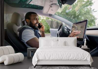 Handsome bearded African man touches the touchscreen in his new high-tech electric vehicle while while talking by phone and smiling. Self driving vehicle concept Wall mural