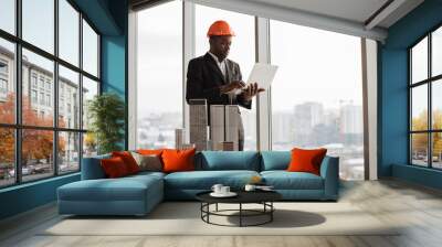 Handsome chief architect african man in business suit and protective helmet with laptop pc working with city buildings model as urban planner at office interior with panoramic city view. Wall mural