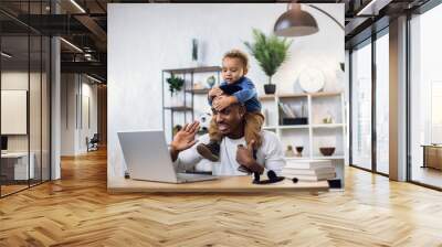 Handsome male freelancer smiling and waving during video chat on modern laptop. Young afro american father working from home and taking care of his little son. Wall mural