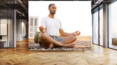 Handsome male meditating in lotus position while sitting on mat on roof of modern house. Young athletic man doing yoga at sunset on urban background. Wall mural