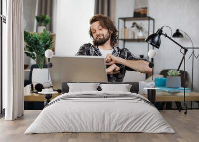 Handsome office man relaxing by stretching his body while sitting in front his computer laptop at the wooden working desk over comfortable living room as background. Wall mural