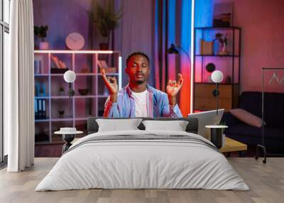 Handsome peaceful young african american man sitting at the desk at home office in the evening and meditating with closed eyes and hands folded in mudra zen gesture Wall mural