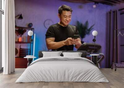 Handsome young sports asian man using phone looking online fitness class while working out, running, doing cardio training on treadmill in evening time, indoor on background of modern apartment Wall mural