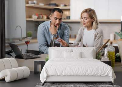 Happy multinational couple using cell phone while having breakfast at kitchen, positive man and woman eating healthy food, checking photos on social media and smiling, having video conference Wall mural