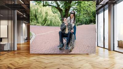 Happy young hipster modern woman with dreadlocks, walking together with her old senior grandfather, sitting in a wheelchair, outdoors in park, talking and laughing. Disabled man with his care giver Wall mural