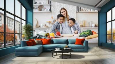 Healthy food, home resting concept. Pretty young mother and two children sons preparing salad with fresh vegetables, hugging and smiling looking at the camera while cooking in kitchen. Wall mural