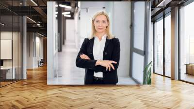 Indoor office portrait of pretty mature blond business lady, wearing white shirt and black jacket, standing with arms crossed in modern office interior. Businesswoman in office corridor Wall mural