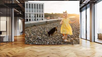 Lively pretty little girl wearing stylish yellow dress playing with dachshund dog on summer terrace of high-rise building. Happy childhood with love for pets. Copy space. Wall mural