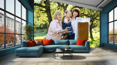 Lovely Caucasian three generation family, grandmother, mother and little cute child kid girl, standing outside in summer spring park and painting a picture on wooden easel. Wall mural