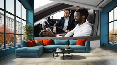 Modern car interior visible, professionalism, customer service, and positive buying experience. Caucasian car dealer sit inside vehicle with African American buyer discussing car features and details. Wall mural