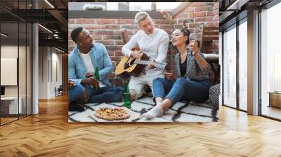 Multi ethnic group of people playing guitar and singing during rooftop party. Young hipsters drinking alcohol and eating pizza. Party time during weekends. Wall mural