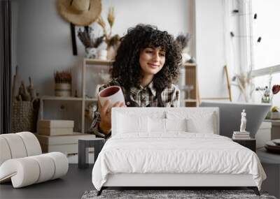 Owner of pottery workshop updates database of items that sold on website, sitting at table against background of shelves with products. Caucasian businesswoman engaged in filling social networks. Wall mural