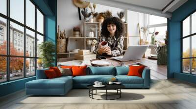 Owner of pottery workshop updates database of items that sold on website, sitting at table against background of shelves with products. Caucasian businesswoman engaged in filling social networks. Wall mural