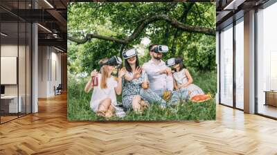 Parents and their two daughters wearing VR headset during picnic at summer garden. Young family having fun with innovative gadgets outdoors. Modern lifestyles. Wall mural